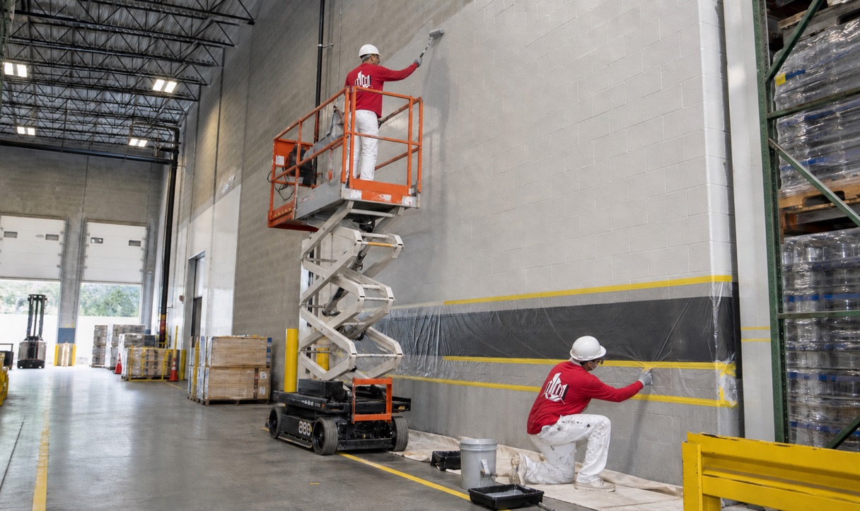 Commercial painting crew coating warehouse walls with lift access and protected inventory